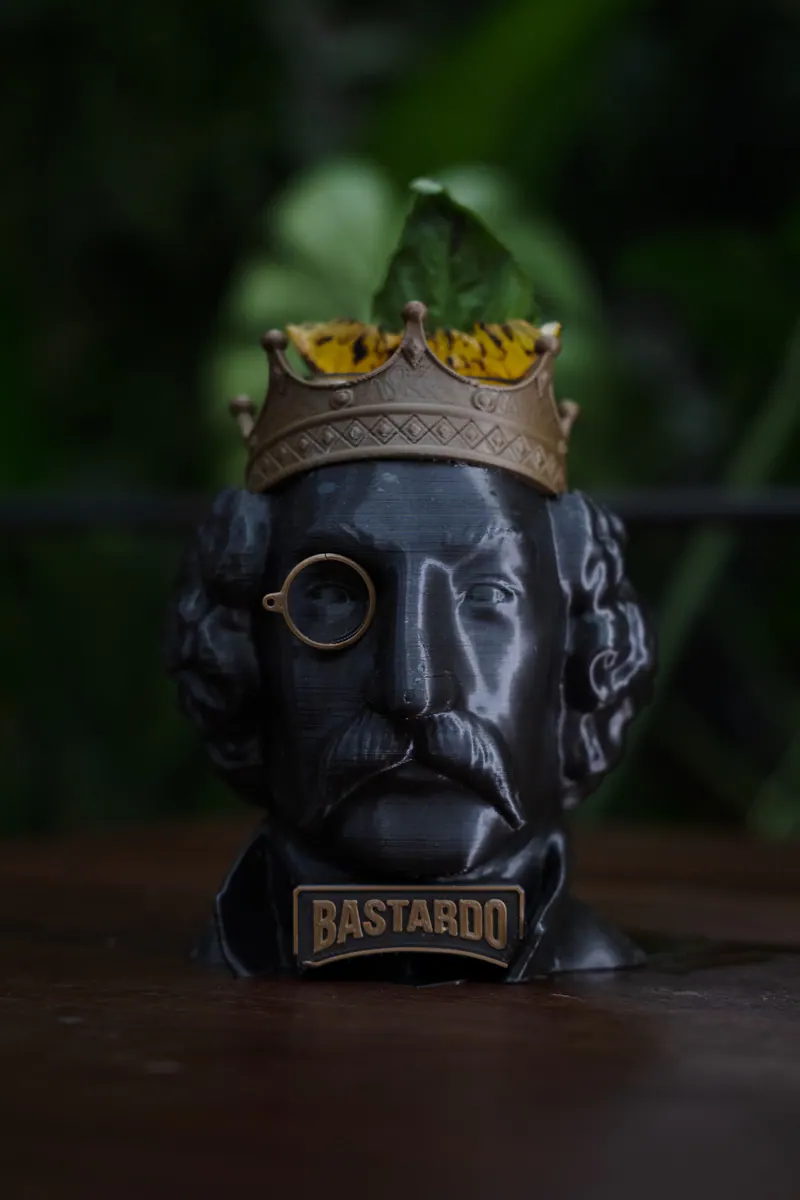 Cóctel Rey Bastardo servido en un busto negro del Rey, con corona de cobre y monóculo, sobre follaje verde