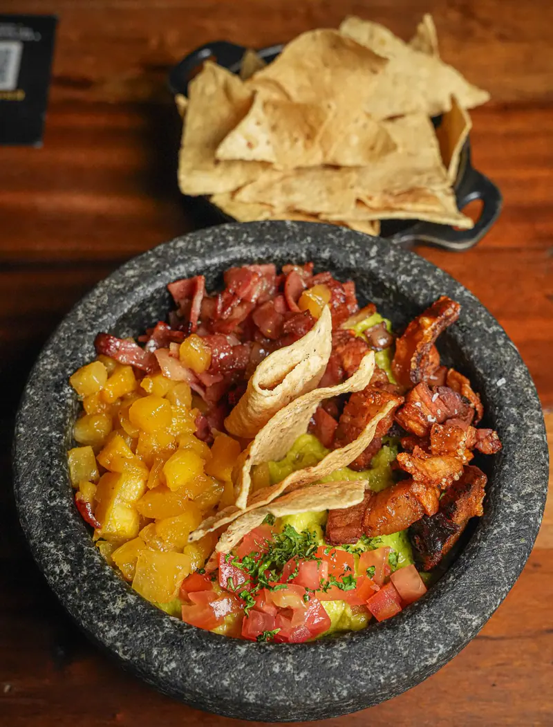 Molcajete de piedra con guacamole, chicharrón, tocineta, tomate y piña asada, con totopos de maíz al fondo