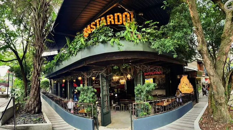 Fachada de Bastardo Envigado: la terraza con el letrero BASTARDO, entre árboles, sobre la Calle de la Buena Mesa